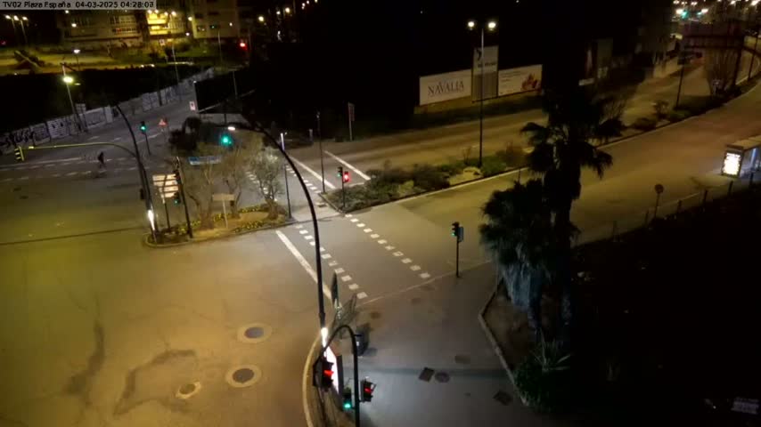 A nighttime, high-angle view shows a mostly empty intersection with traffic lights and palm trees, under well-lit conditions.