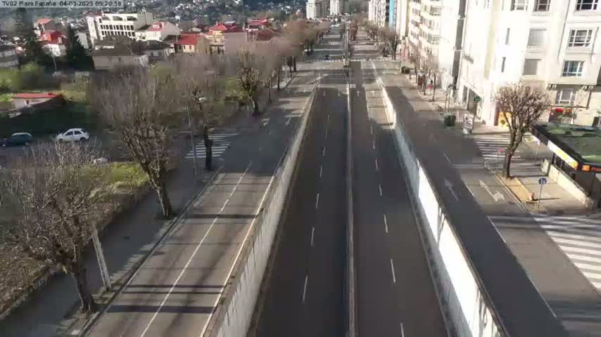 A mostly empty, two-lane road runs through a city on a sunny day, flanked by buildings and bare trees.