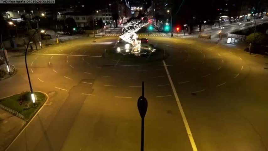 A nighttime aerial view of a mostly empty roundabout with a light sculpture in the center, illuminated by streetlights.