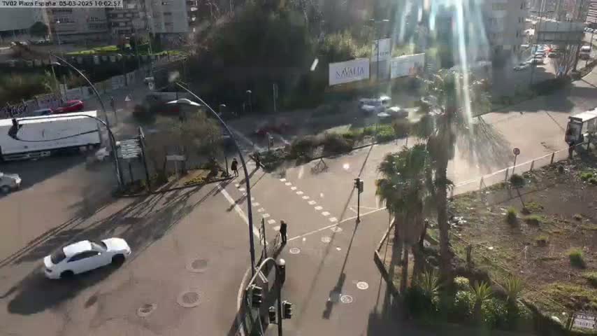 A sunny day reveals a high-angle view of a city intersection with several cars, a truck, and pedestrians, along with palm trees and buildings in the background.