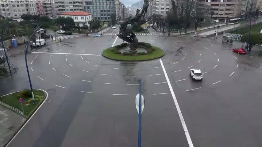 A wet roundabout with a large metal sculpture in the center, surrounded by buildings and a few cars on a rainy day.