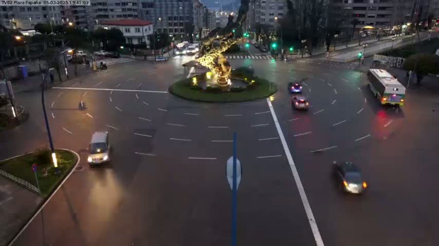 A roundabout at night with several cars and a bus navigating it under dark, possibly wet conditions.