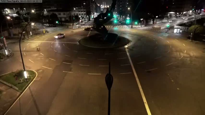 A nighttime aerial view shows a mostly empty roundabout with a dark sculpture in the center, surrounded by city streets and buildings.