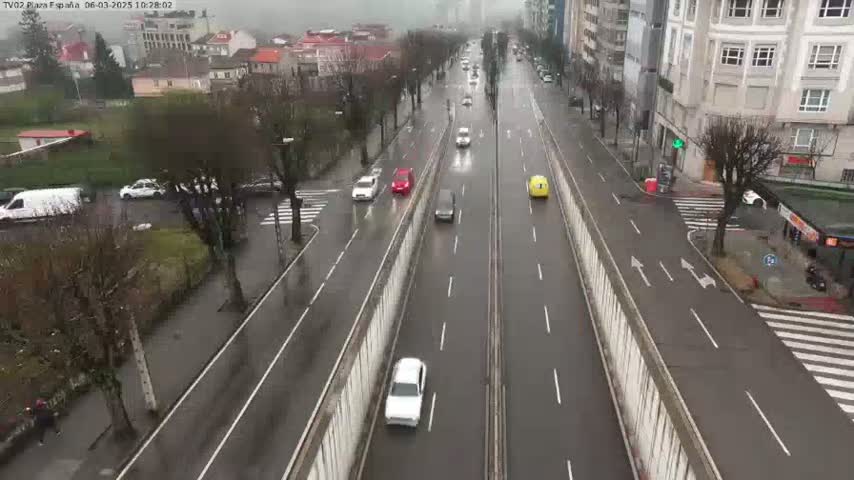 A wet, multi-lane road with several cars driving in both directions, bordered by buildings and trees on a rainy day.