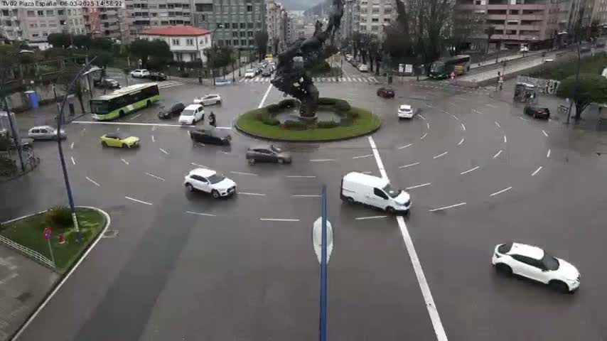 Several cars and a bus navigate a wet roundabout surrounding a central sculpture in a city setting.