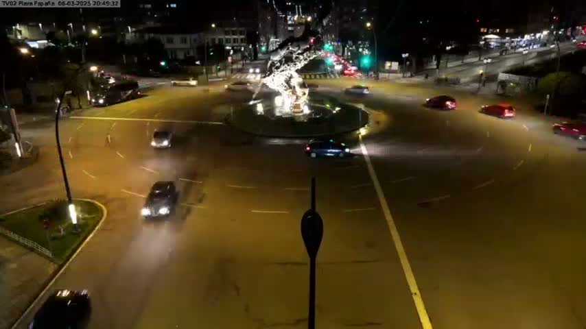 A nighttime aerial view shows several cars navigating a roundabout centered on a light-emitting fountain, with some streetlights illuminating the wet asphalt roads.