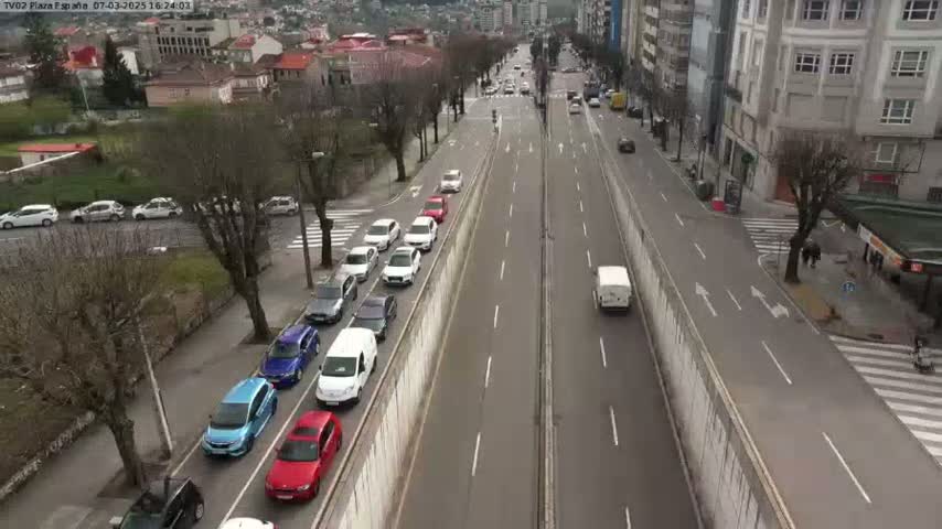 A long, two-lane road with many cars parked along a wall and cars driving in both directions on a mostly clear day.
