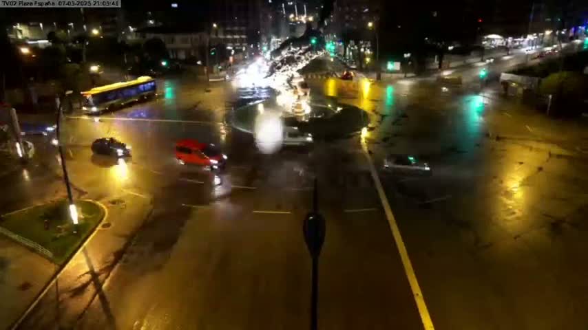 A wet, rain-slicked city intersection at night is illuminated by streetlights, showing several vehicles and a fountain in the center.