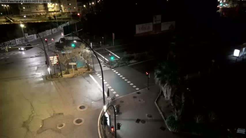 A nighttime, overhead view of a mostly empty intersection with streetlights illuminating wet roads and surrounding buildings.