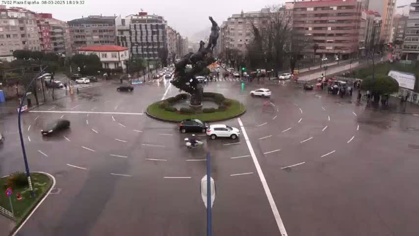 A wet roundabout in a city, featuring a large bronze sculpture in the center, surrounded by several cars and pedestrians under a rainy sky.