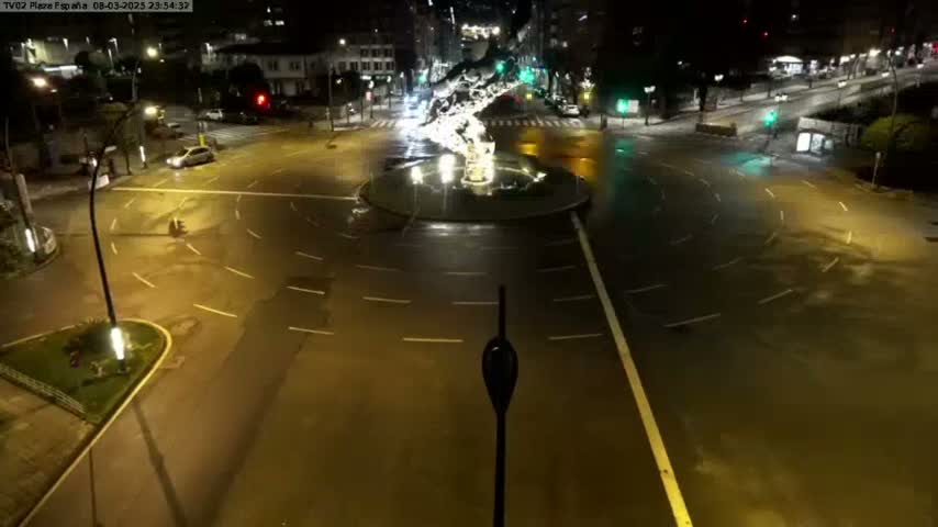 A wet, nighttime roundabout with minimal traffic is illuminated by streetlights.