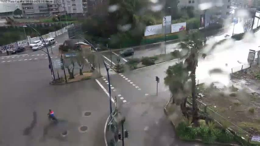 A rainy day scene shows a street intersection with several cars and a person on a bicycle; heavy rain is obscuring visibility.