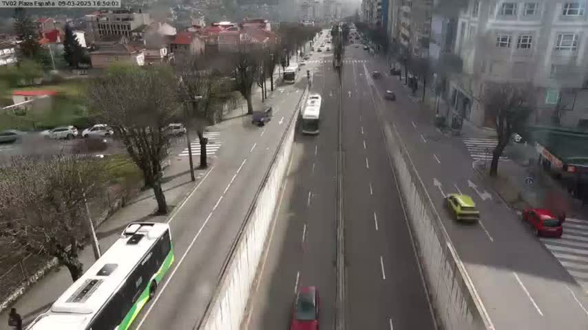 A high-angle view shows a city street with several cars and buses moving in both directions, alongside a tall concrete barrier, under an overcast sky.