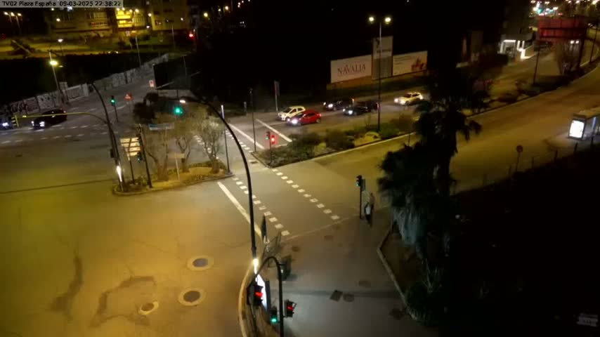 A nighttime aerial view of a multi-lane intersection with several cars, illuminated by streetlights, with palm trees and other landscaping visible.