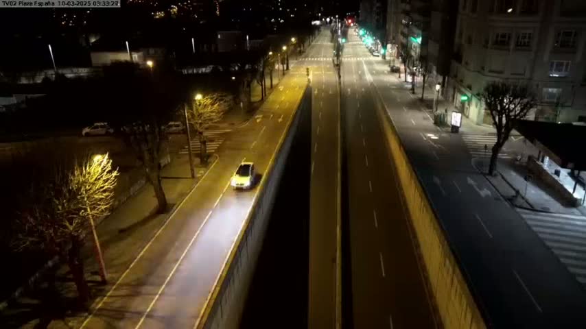 A mostly empty, well-lit city street at night, with a single car driving on one side, is viewed from above.