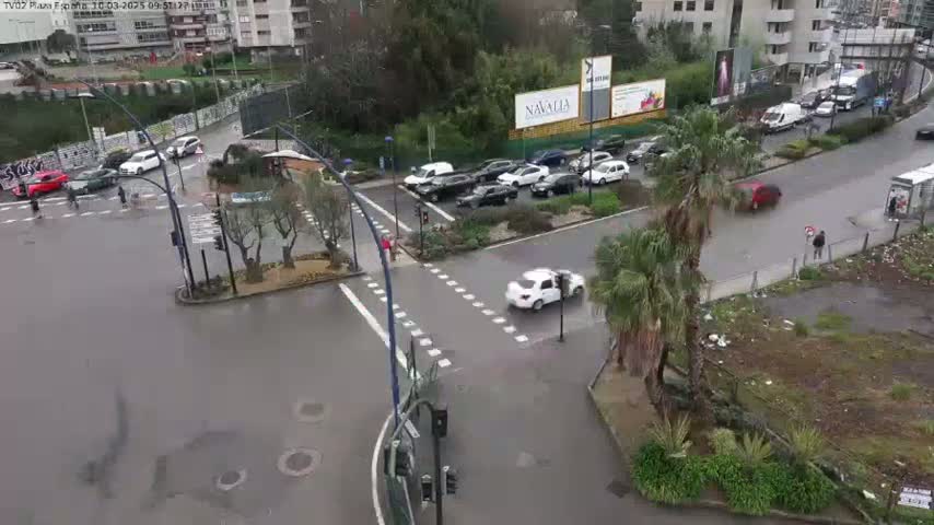 A rain-slicked city intersection shows several cars navigating a busy roadway, with pedestrians and some greenery visible near the corners.
