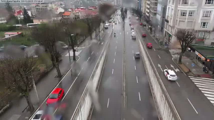 A rain-slicked city street with multiple lanes of traffic, flanked by buildings and trees.