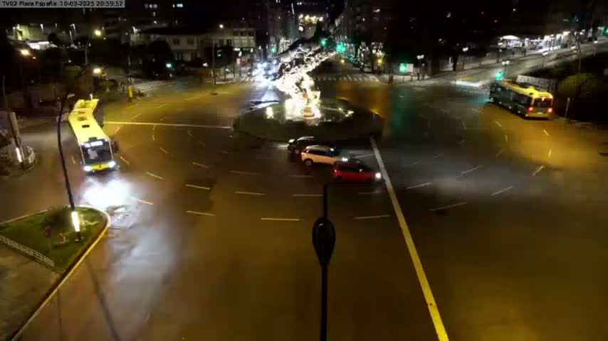 A wet, nighttime roundabout with several cars and buses in motion around a central fountain.