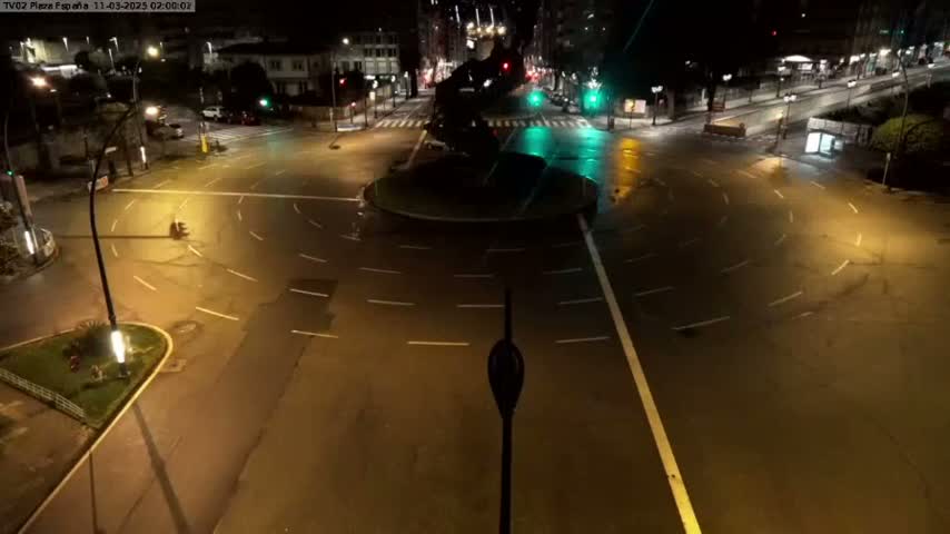 A wet, empty roundabout at night, illuminated by streetlights, is surrounded by dark buildings and roads.