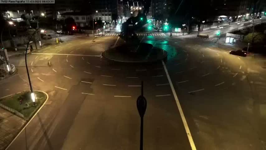 A nighttime aerial view of a mostly empty roundabout with several cars, illuminated by streetlights.