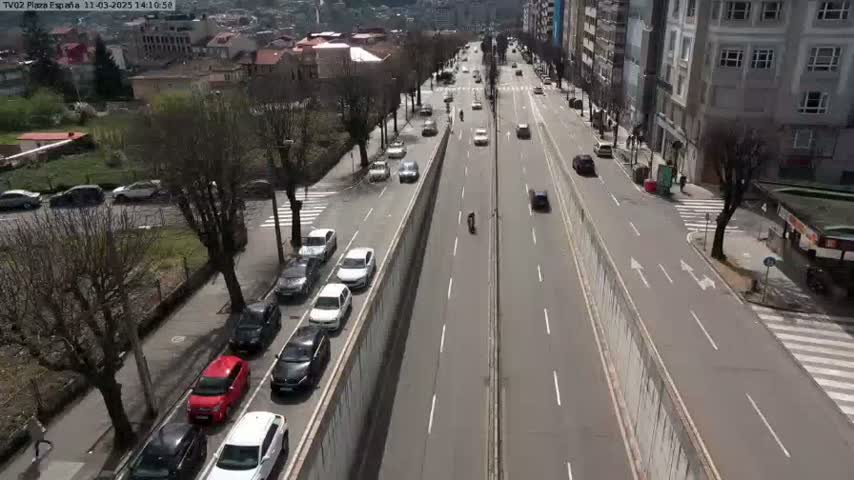 A high-angle view of a multi-lane road with cars driving in both directions, lined with buildings and trees on a sunny day.
