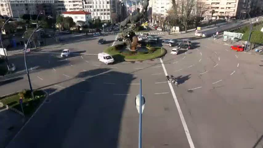A sunny day shows a roundabout with several cars and motorcycles, surrounded by city buildings.