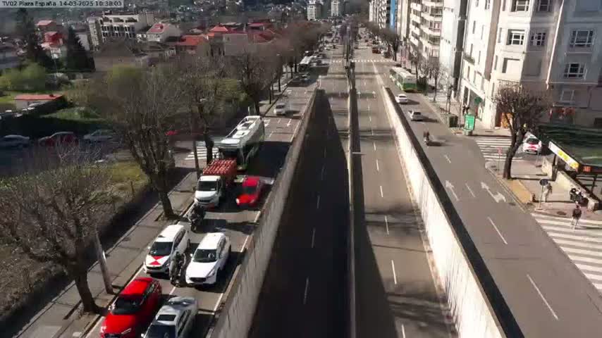 A sunny day reveals a high-angle view of a city street with multiple vehicles, including cars and buses, traveling in both directions alongside a tall concrete barrier separating the roadways.