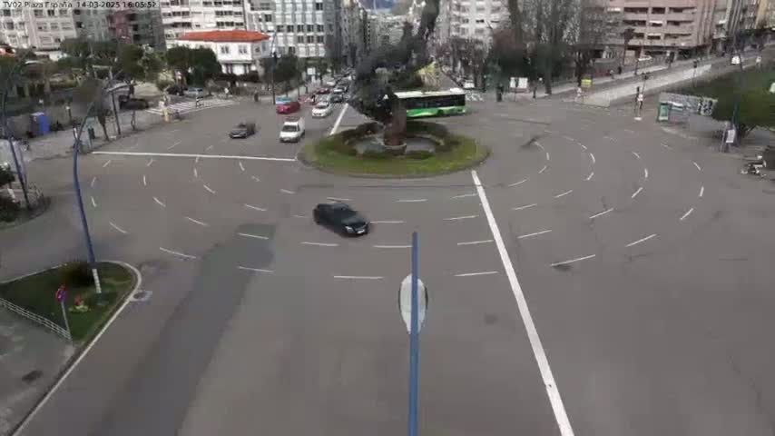 A roundabout with several cars driving around a central statue and surrounded by buildings on a mostly clear day.
