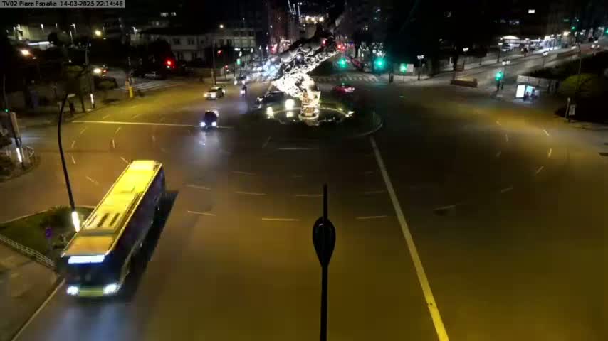 A nighttime aerial view shows a roundabout with several vehicles, including a long bus, and streetlights illuminating the wet roadway.