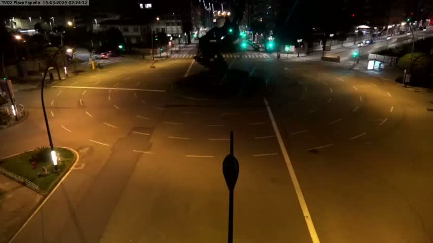 A mostly empty roundabout at night is illuminated by streetlights, with a few vehicles visible in the distance.