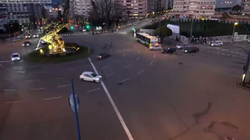 A high-angle, nighttime view of a roundabout with several cars and a bus, under dim lighting conditions.