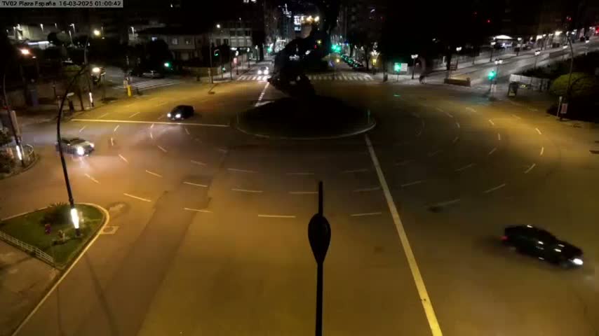 A nighttime aerial view shows a mostly empty roundabout with a few cars driving around it on wet asphalt under bright streetlights.