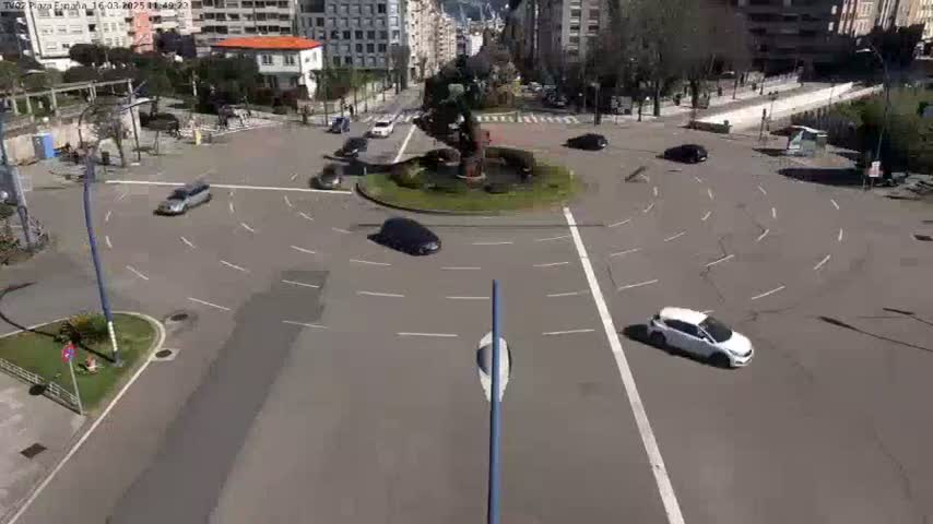 A mostly empty roundabout with several cars driving around a central fountain, on a sunny day.