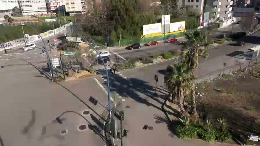 A sunny aerial view of a city intersection with several cars and pedestrians, palm trees lining the streets, and buildings in the background.