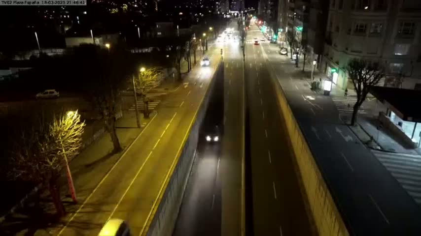 An aerial nighttime view shows a multi-lane road with sparse traffic, bordered by buildings and trees under streetlights.