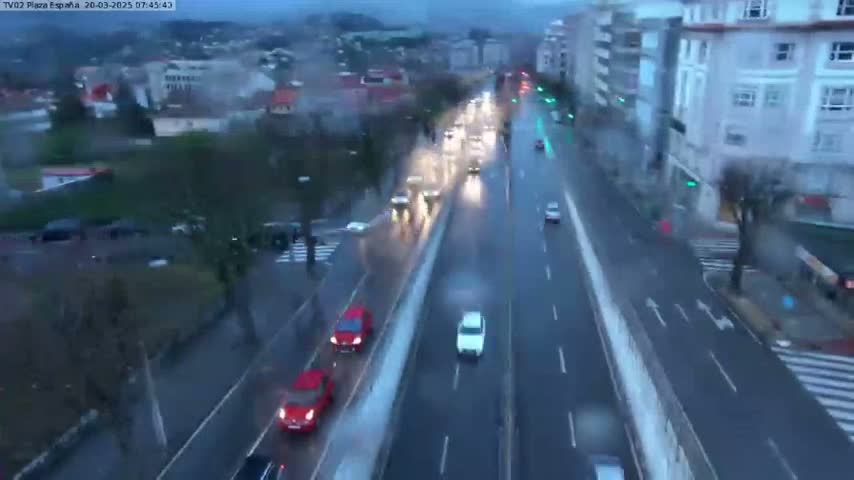 A rainy day view of a multi-lane road with several cars driving in both directions, alongside buildings and greenery.