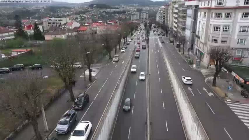 A multi-lane road with moderate traffic flows through a city,  surrounded by buildings and trees on an overcast day.