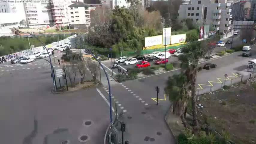 A high-angle, daytime view of a city intersection with several cars stopped and moving, surrounded by buildings and greenery under a clear sky.