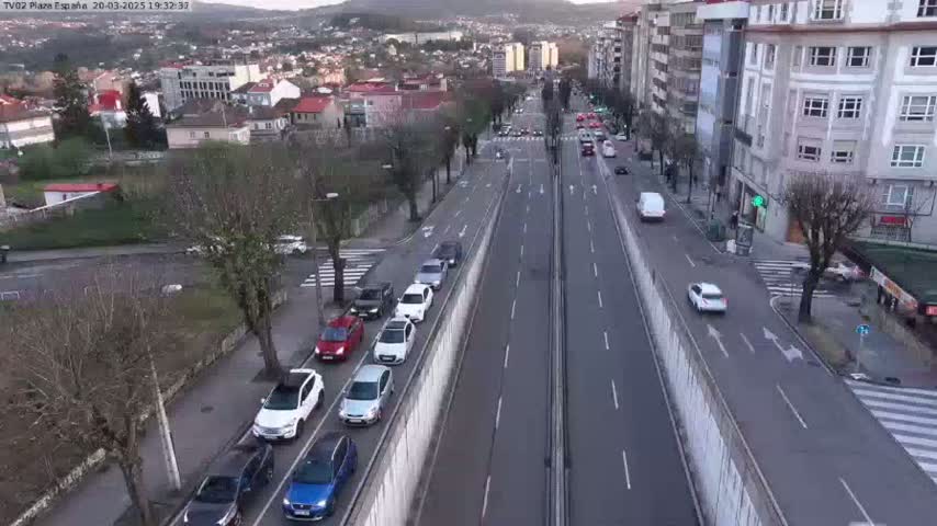 A slightly overcast day reveals a multi-lane road with moderate traffic flowing in both directions, flanked by buildings and trees.