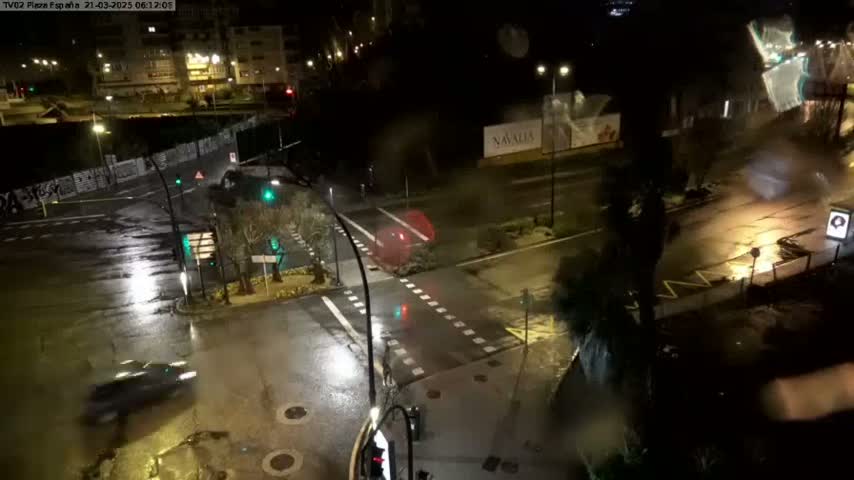 A rainy night scene shows a wet intersection with several cars, streetlights, and buildings in the background.