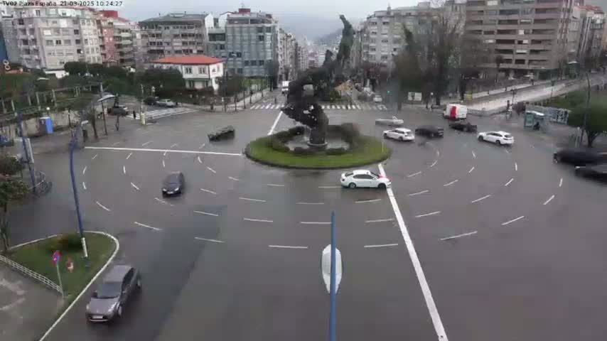 A wet roundabout in a city, surrounded by buildings, with several cars navigating the circular lanes.