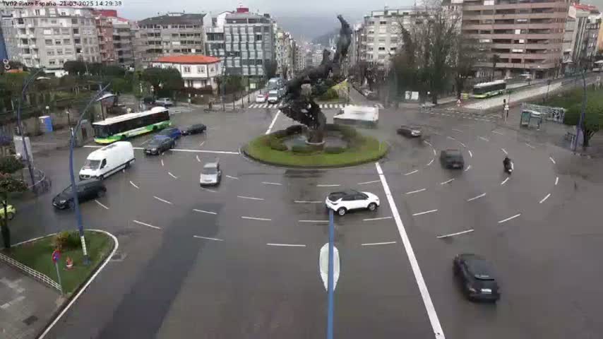 A wet roundabout with several cars and a bus, surrounded by buildings, features a large metal sculpture in the center.