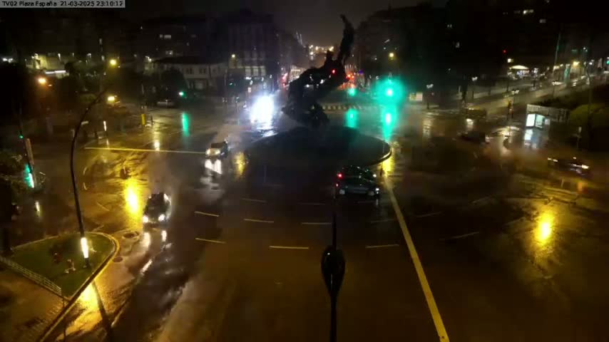 A nighttime view of a rain-soaked roundabout with a large statue in the center, surrounded by cars and streetlights.