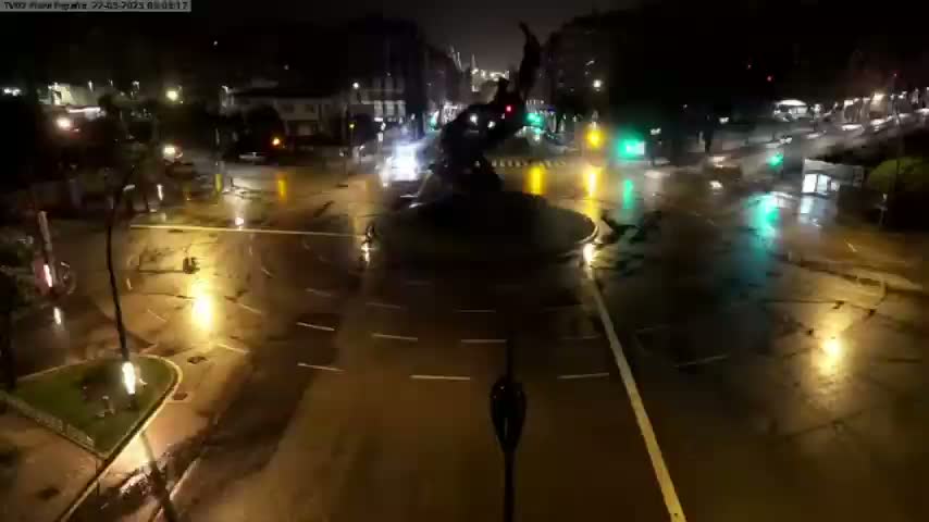 A wet, nighttime street scene shows a roundabout with a dark statue at its center, surrounded by sparsely trafficked roadways with streetlights reflecting on the puddles.
