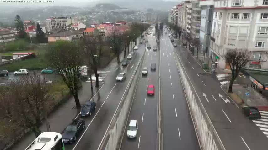 A wet, city street with several cars driving in both directions, surrounded by buildings and trees.