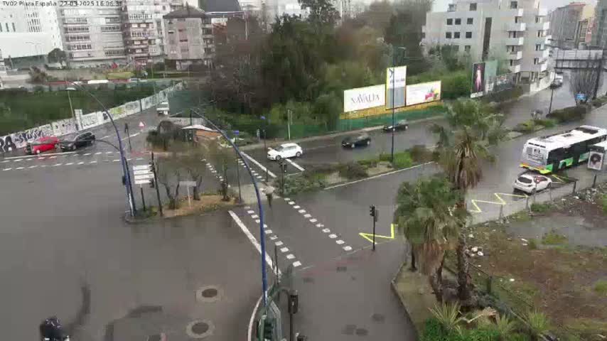 A wet, city intersection with several cars and a bus, surrounded by buildings and palm trees.