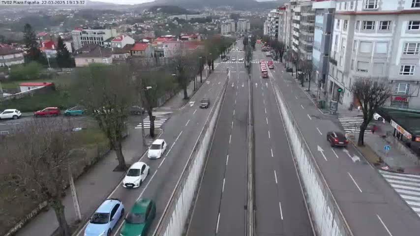 A wide, divided highway with several cars driving on it, flanked by buildings and trees under an overcast sky.
