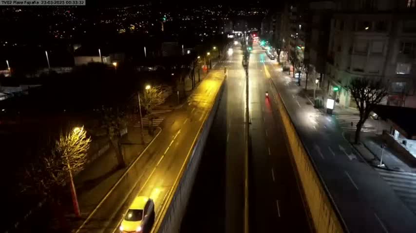 A mostly empty, well-lit city street at night, with a few cars visible, viewed from an elevated perspective.