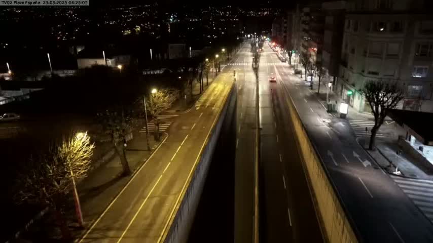 A wet, empty road at night is lit by streetlights, flanked by buildings and trees, with a few distant cars visible.