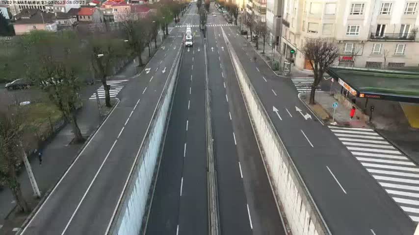 A mostly empty, multi-lane roadway cuts through a city on a cloudy day, with buildings and sparse pedestrian activity visible on either side.